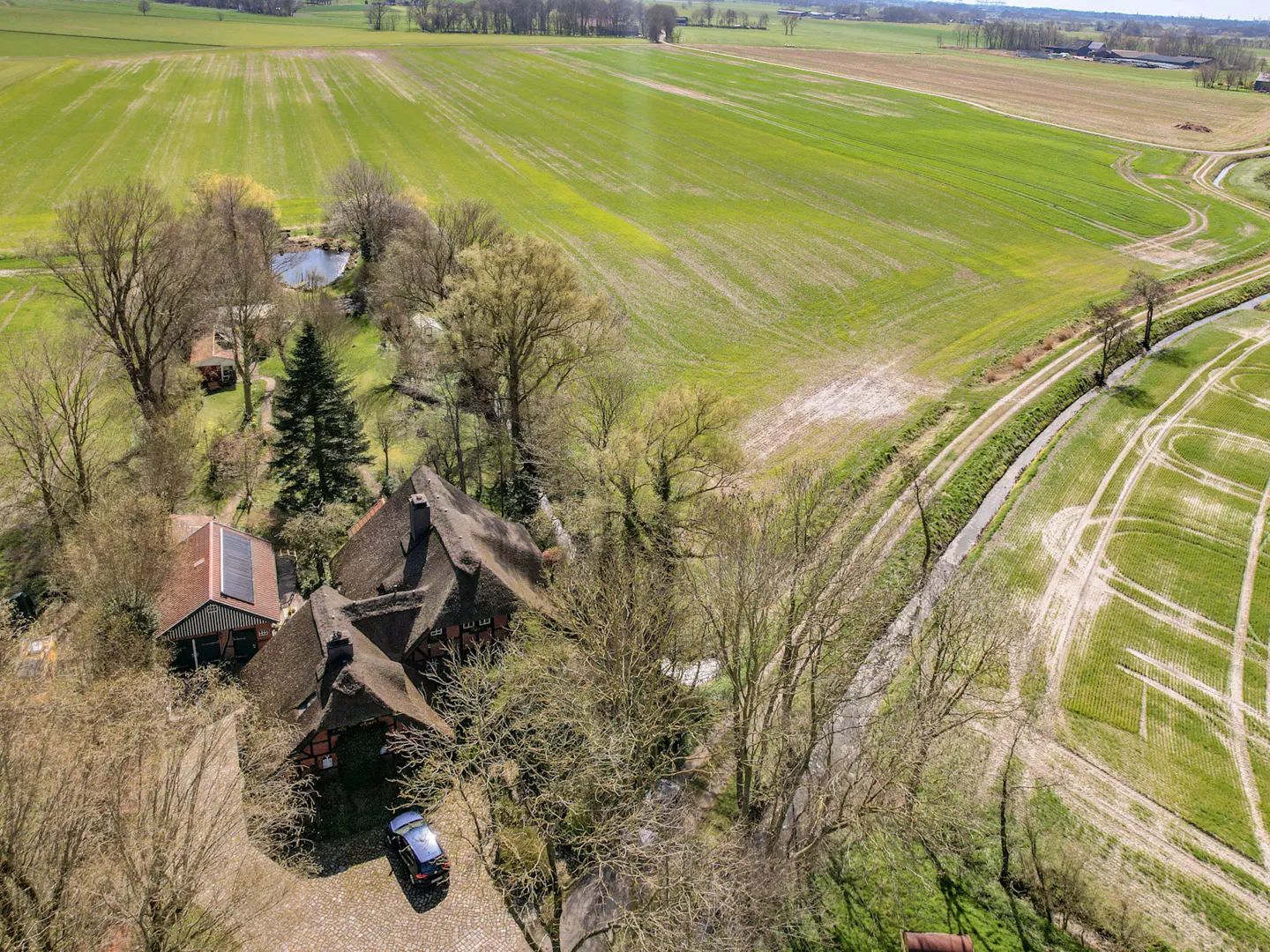 Aerial view of a thatched-roof house with solar panels, surrounded by trees and green fields. A car is parked in the driveway.