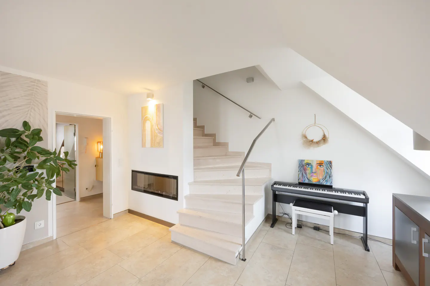 Bright, modern living room with white walls, stairs, fireplace, piano, and artwork. A plant sits in the foreground.