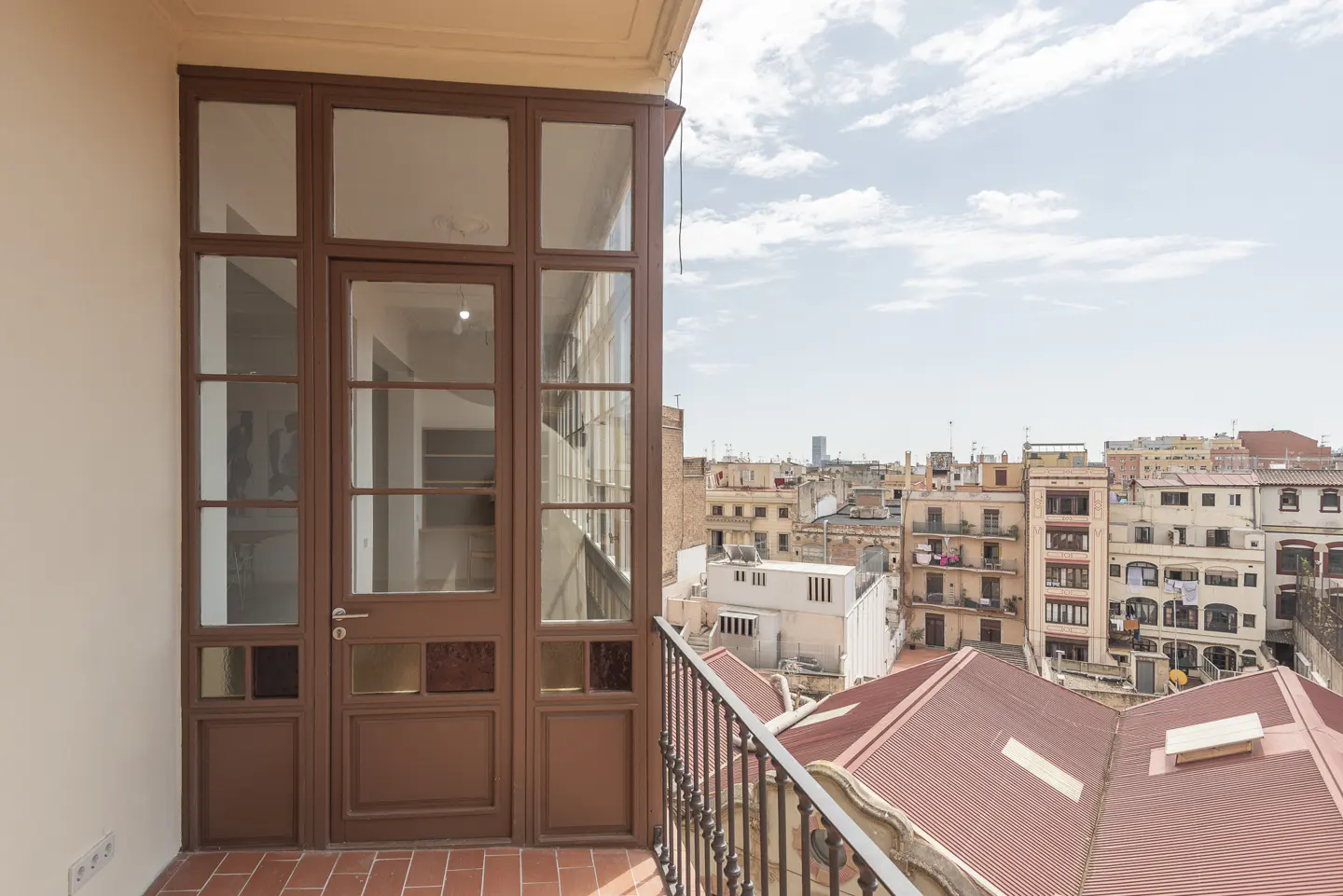 Balcony with brown framed glass doors, red tile floor, and black railing overlooking a cityscape under a cloudy sky.
