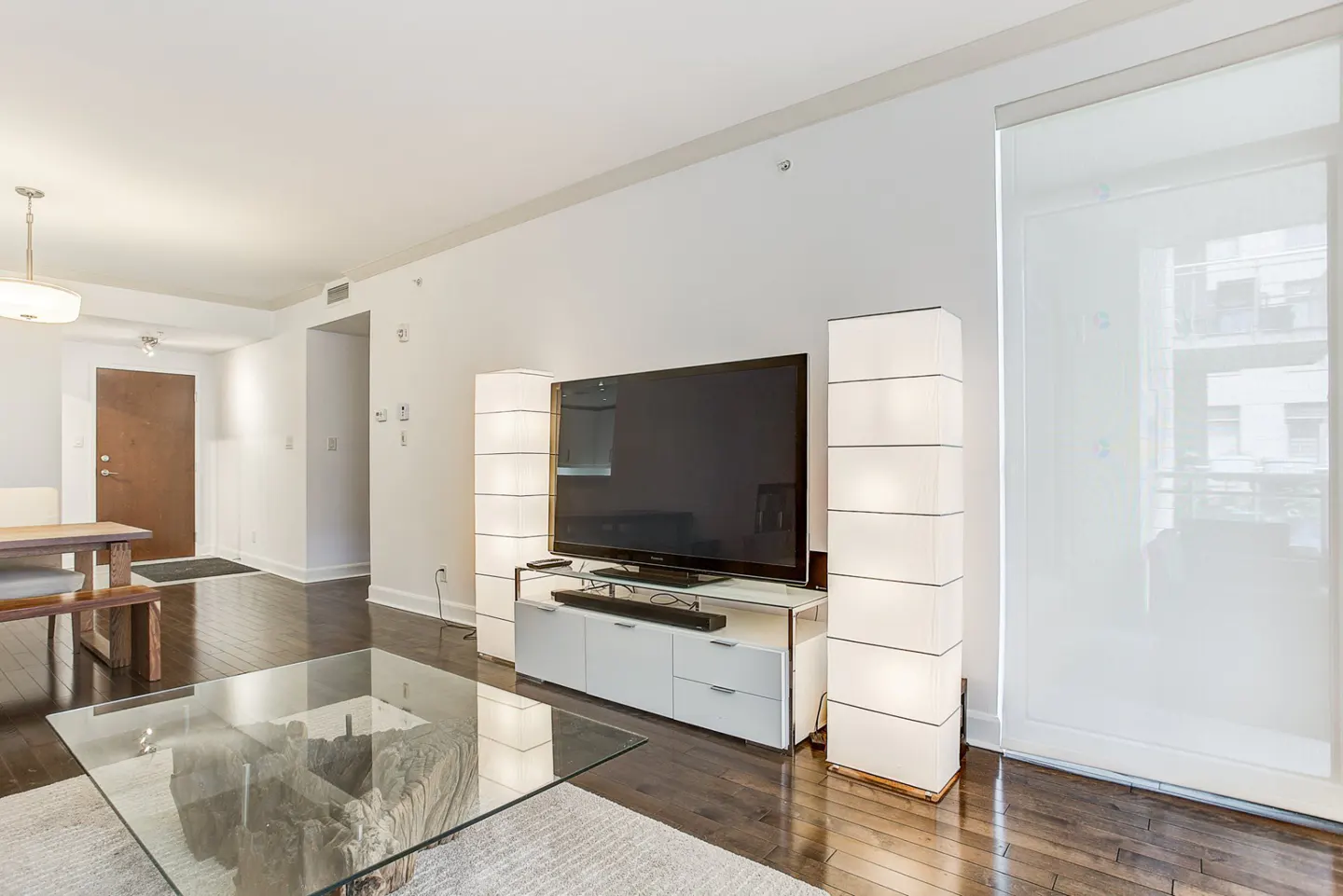 Bright living room with dark wood floors, white walls, and a large TV flanked by two tall, white lamps. A glass coffee table sits on a light rug.