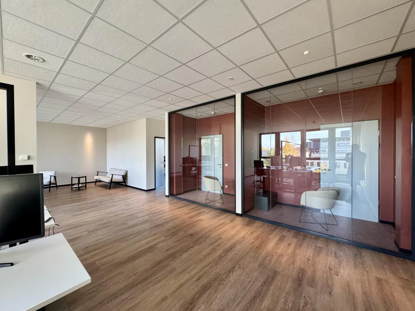 Office space with wood floors, white walls, and glass-walled offices with rust-colored walls. A waiting area has a small sofa and chairs.