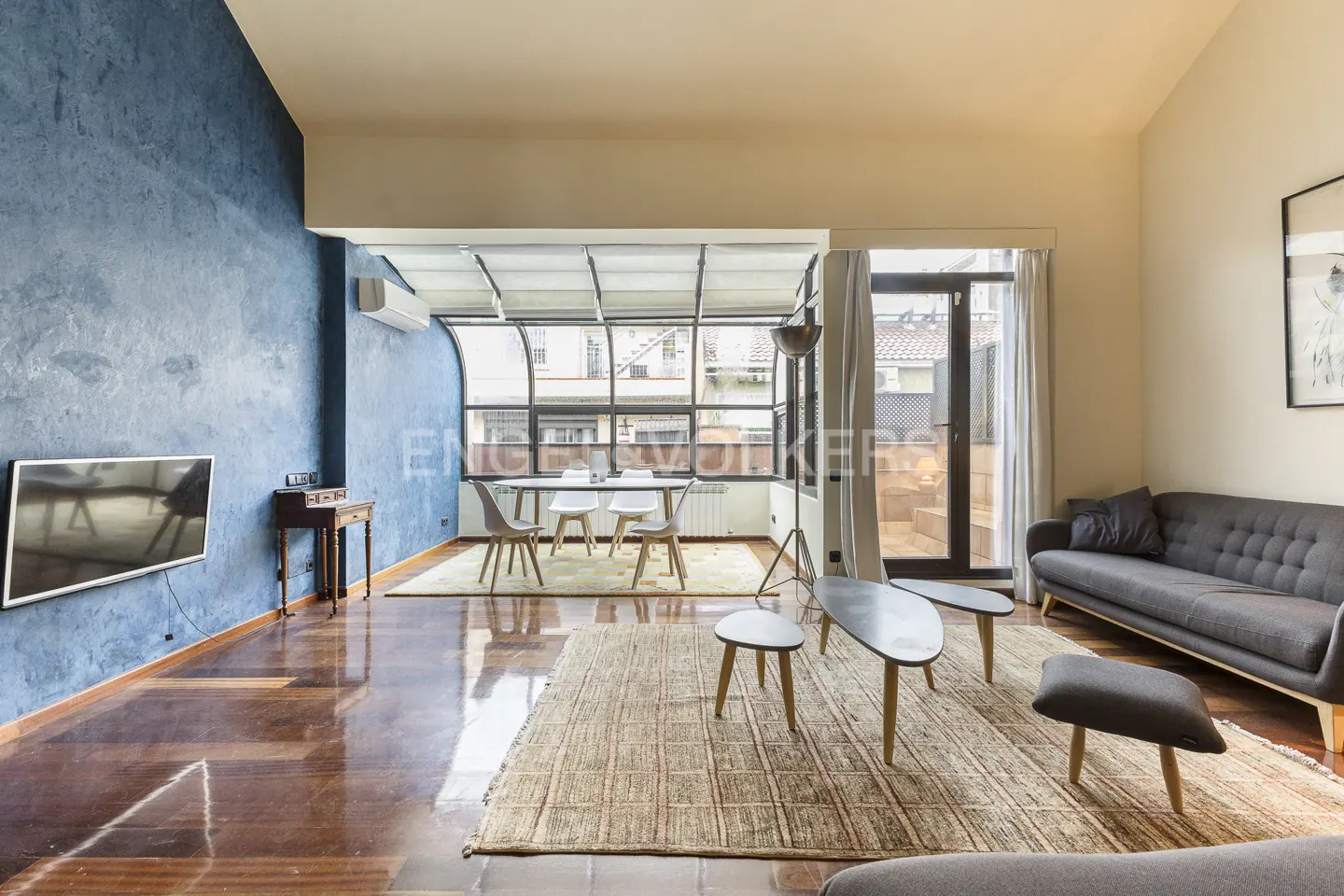 A living room with blue accent wall, hardwood floors, and a dining area with a glass ceiling. A gray sofa and coffee tables sit on a woven rug.