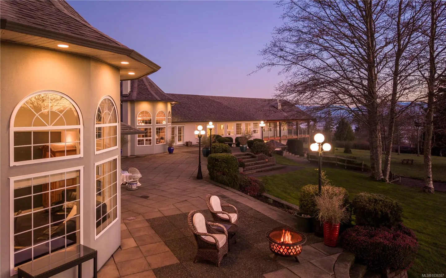 Twilight view of a large home's patio with wicker chairs, a fire pit, and manicured landscaping leading to a covered porch.