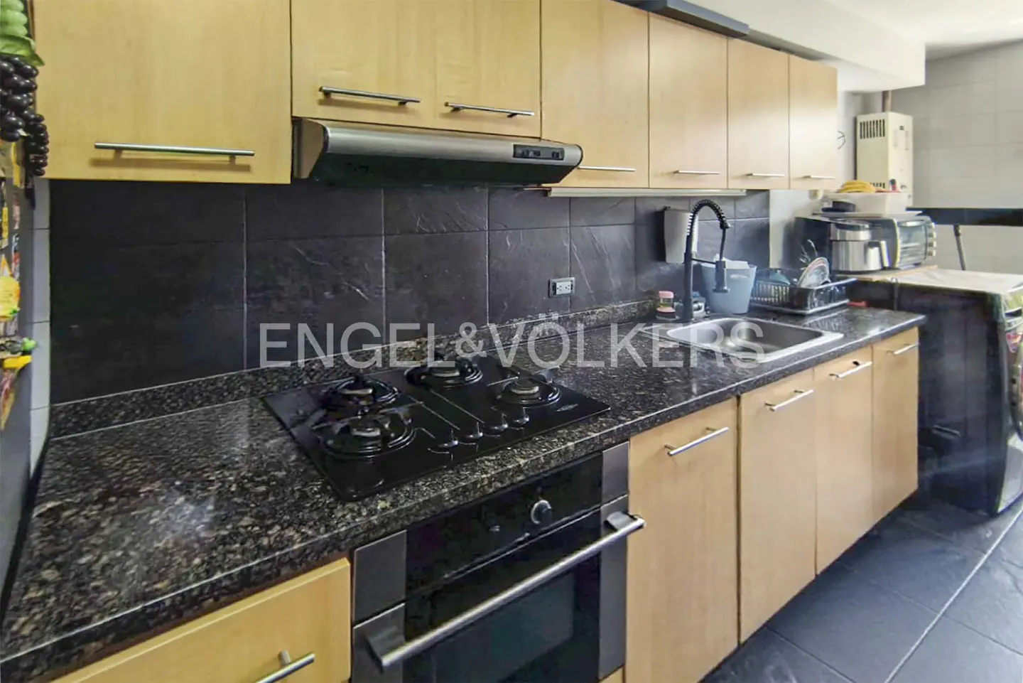 A modern kitchen with light wood cabinets, black granite countertops, and a black backsplash. A gas stovetop and oven are visible.