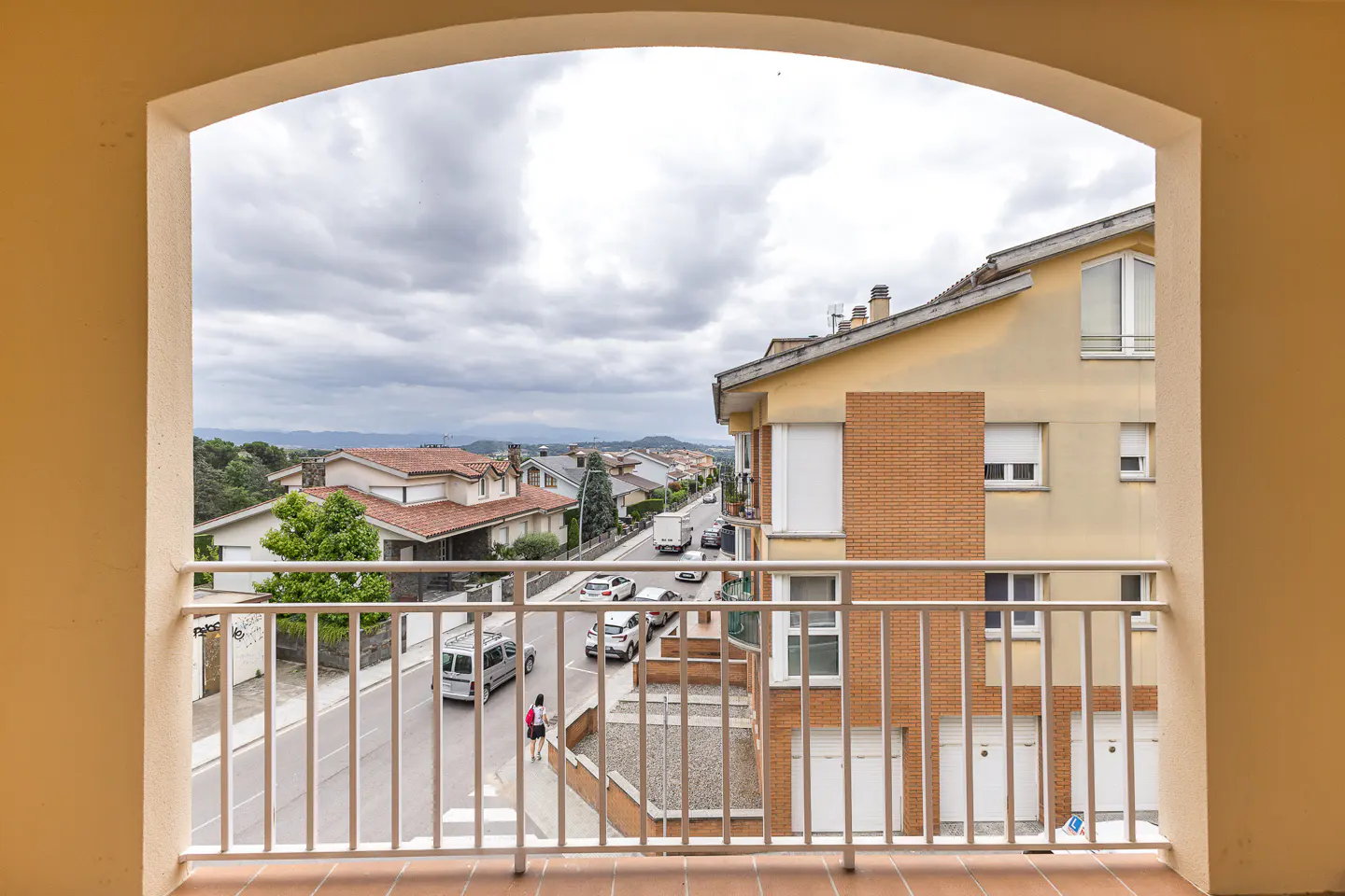 View from a balcony overlooking a suburban street with cars, houses, and a cloudy sky.