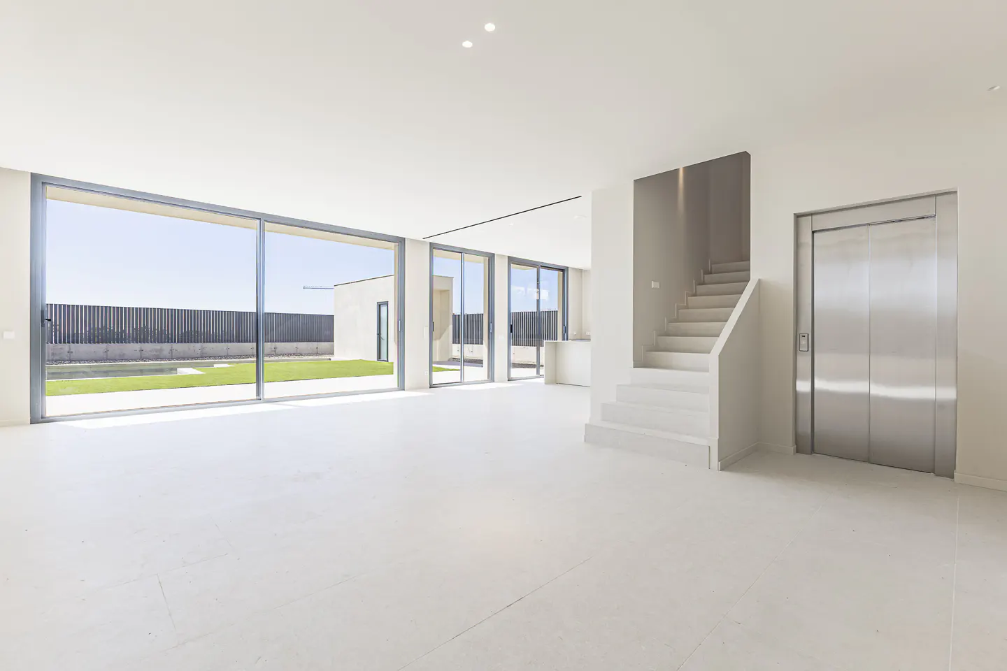 Bright, modern interior with white floors, large sliding glass doors to a green lawn, stairs, and a silver elevator.