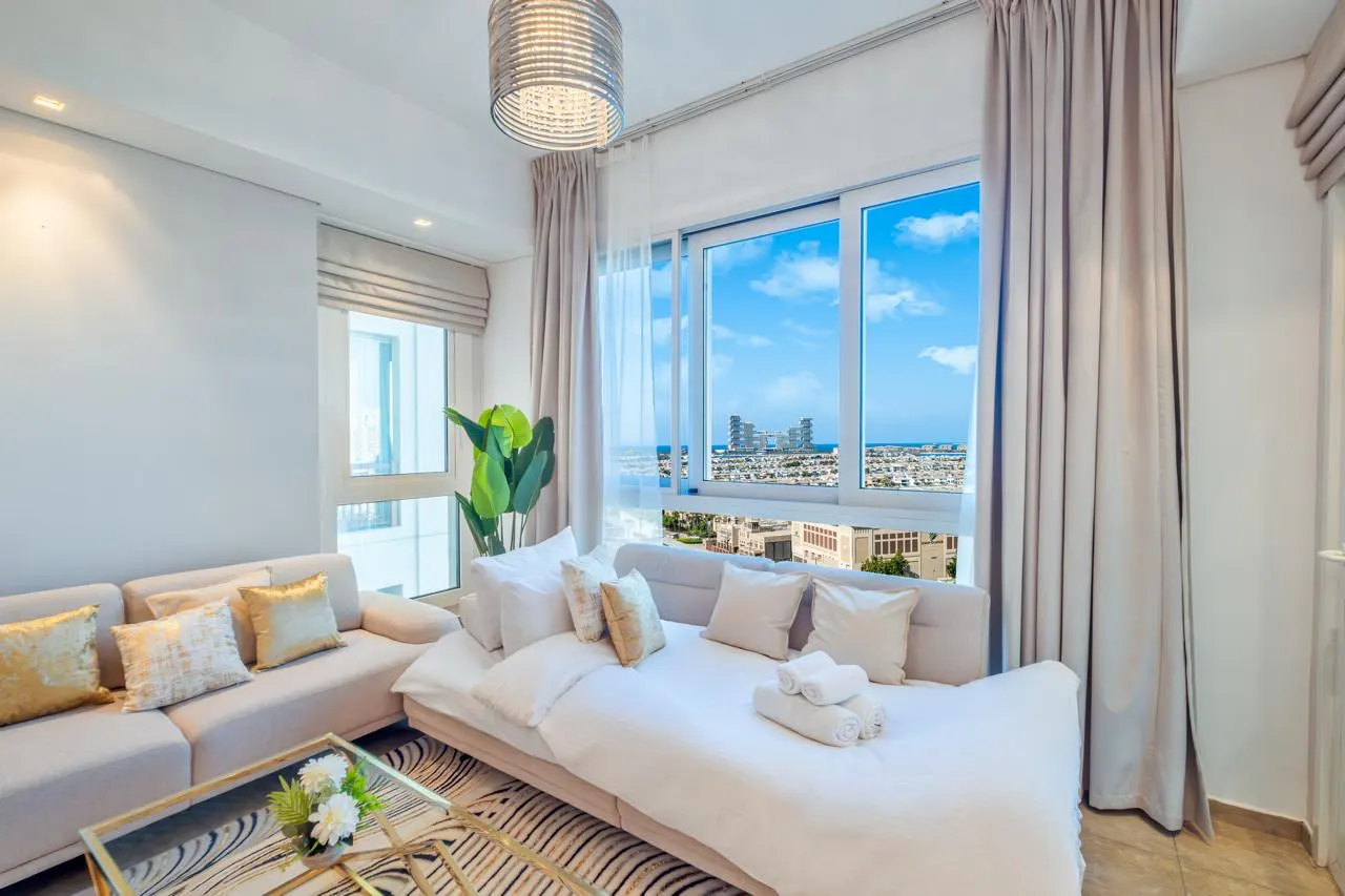 Bright living room with beige sofa, white cushions, and a glass coffee table. Large windows offer a city view with blue sky.