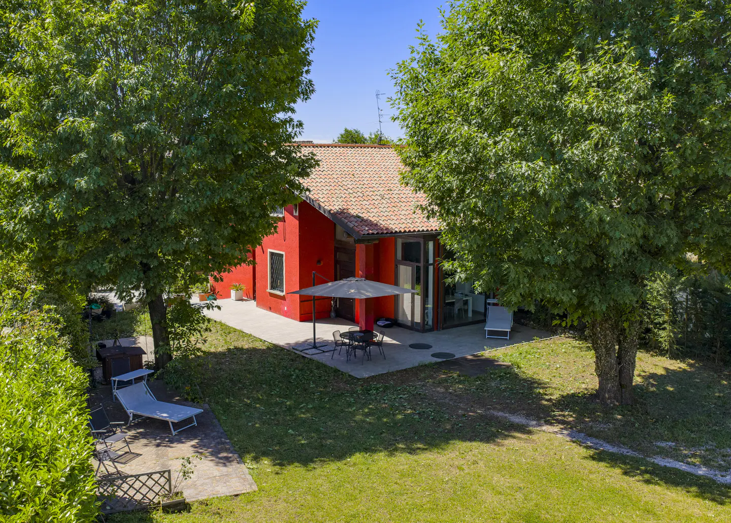 Aerial view of a red house with a terracotta roof, surrounded by green trees and a lawn. Patio furniture sits outside.