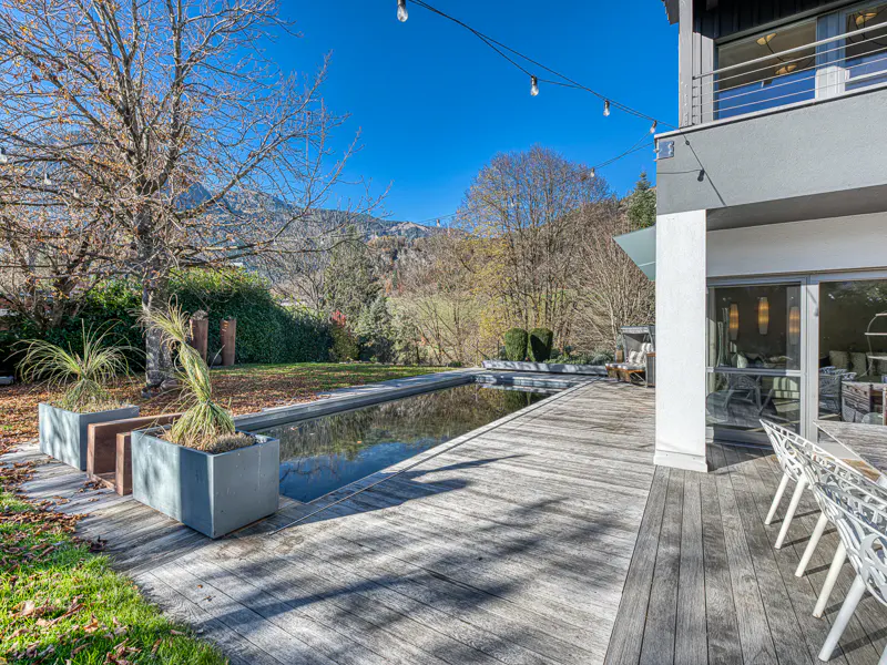 Outdoor view of a modern home with a pool, wooden deck, and mountain backdrop under a clear blue sky. String lights hang above.