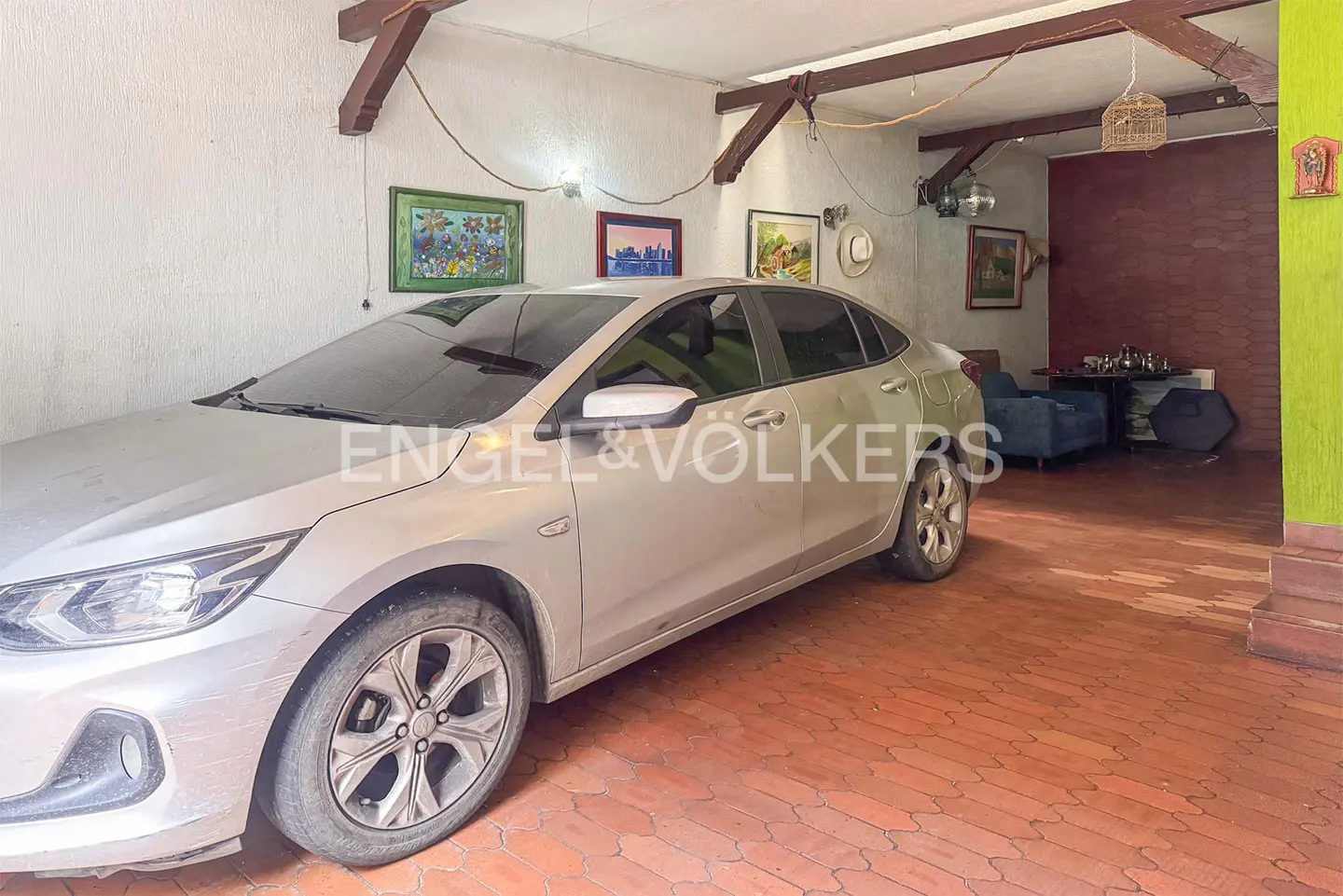 Indoor garage view with a silver car parked on a red brick floor, paintings on the wall, and a living room area in the background.
