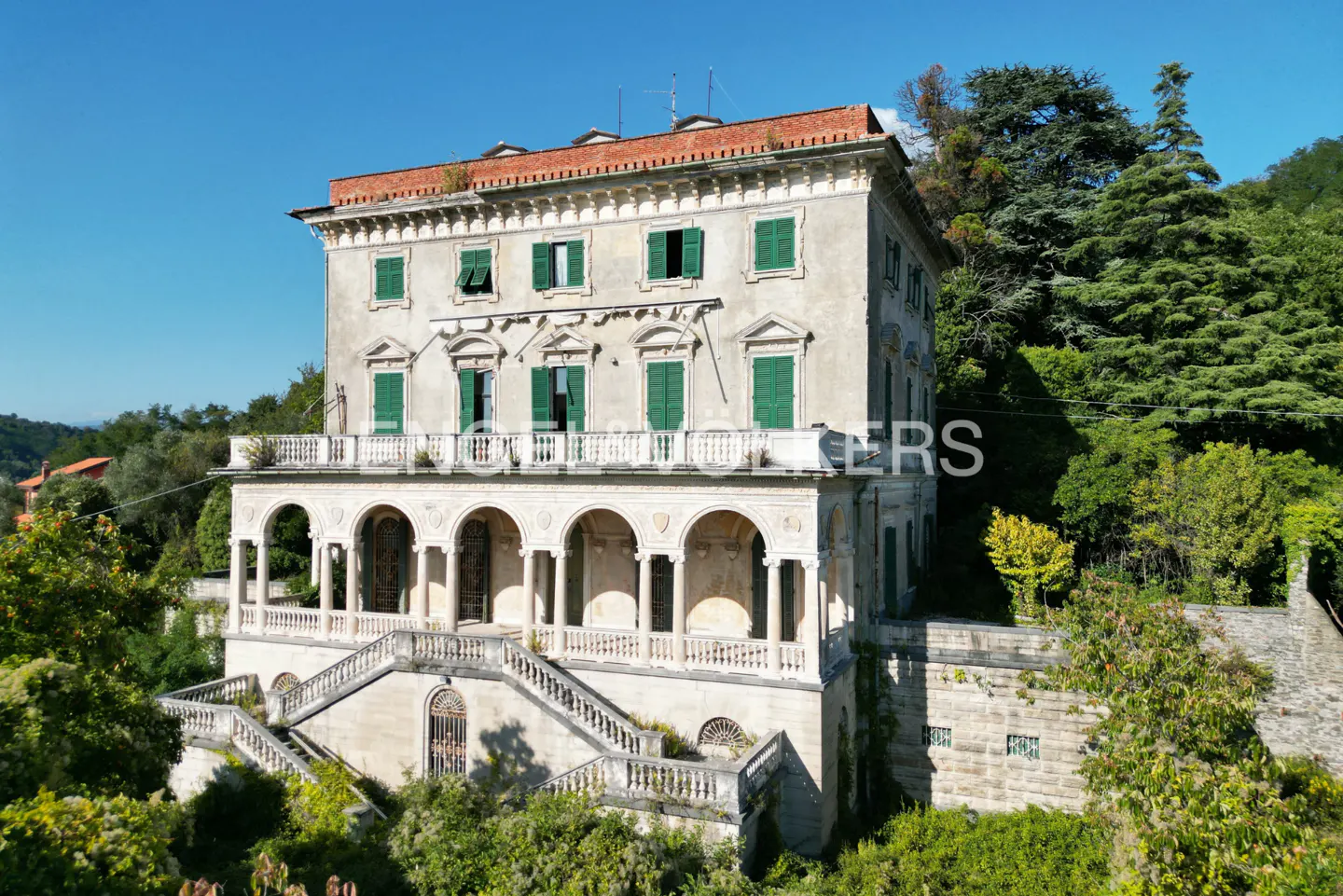 Exterior view of a three-story villa with green shutters, a red tile roof, and a stone staircase.