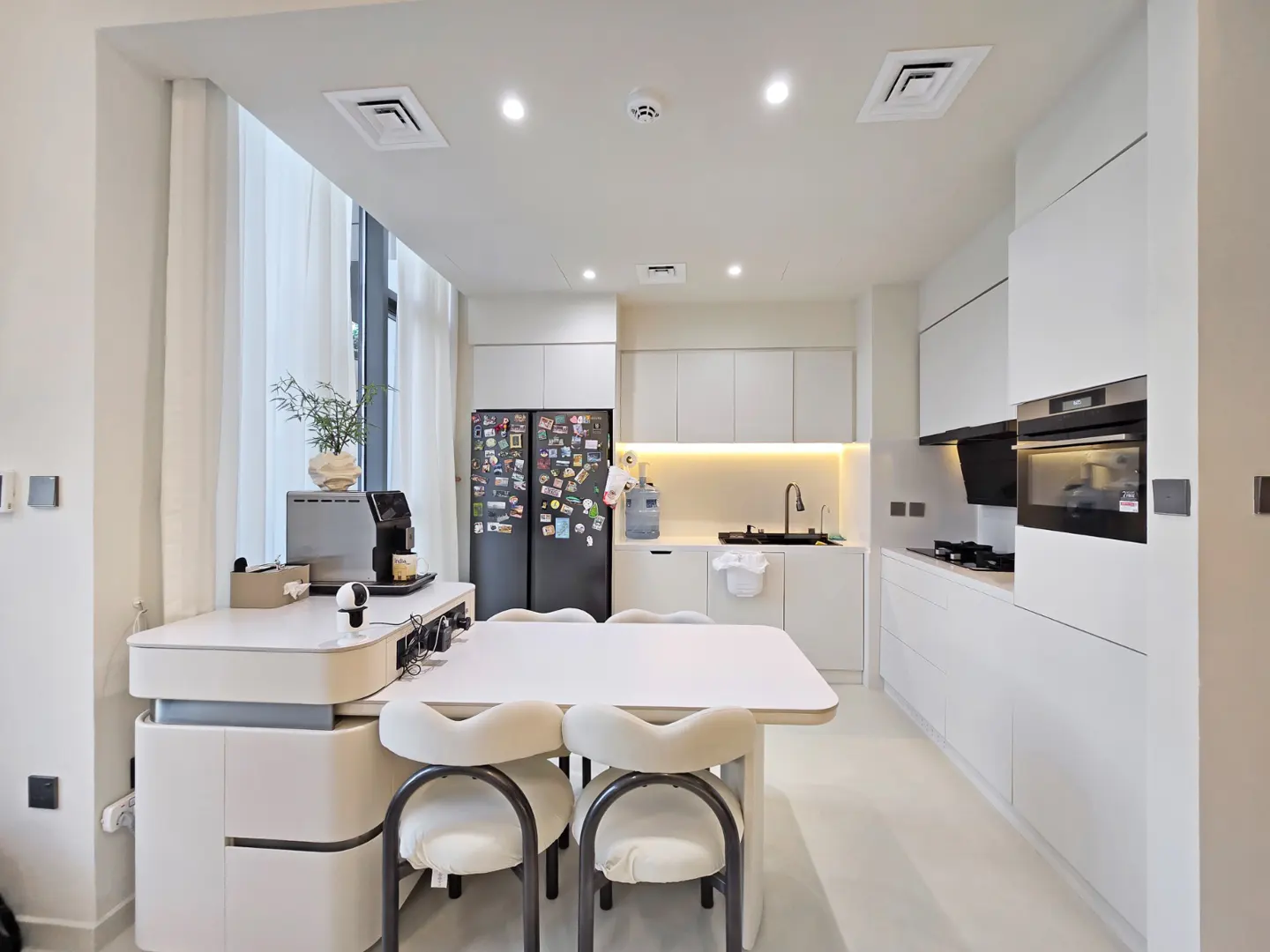 Bright, modern kitchen with white cabinets, black fridge covered in magnets, and a white table with six chairs. Natural light filters through sheer curtains.