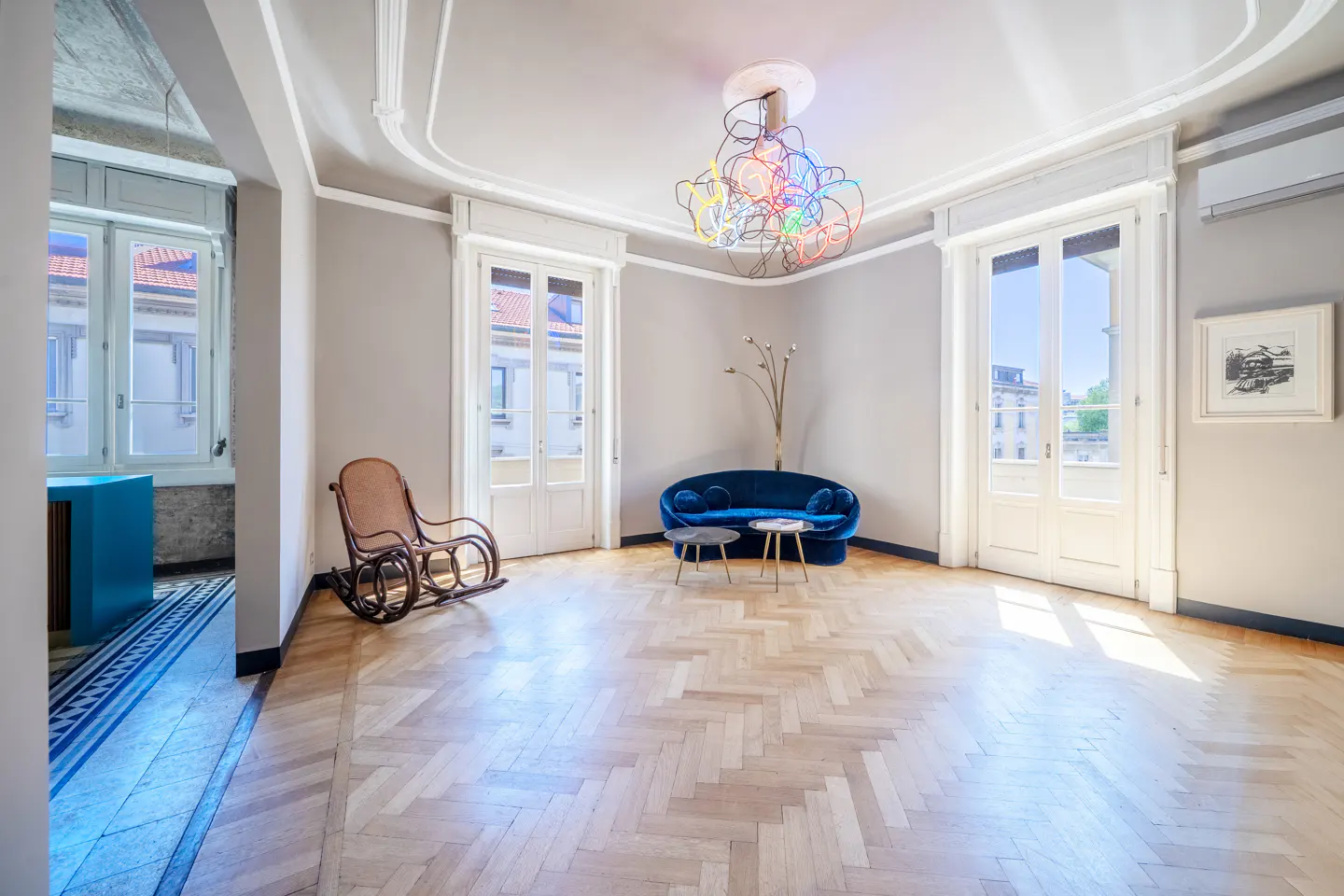 Bright, airy room with herringbone wood floors, a blue velvet sofa, and a colorful modern chandelier. A rocking chair sits near an open doorway.