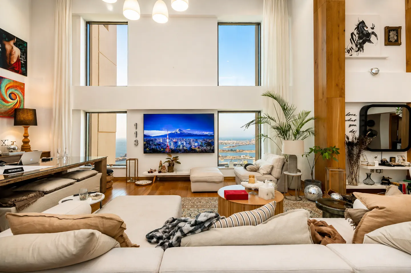 Bright, modern living room with white walls, large windows, and a view of the city and ocean. A large TV displays a mountain scene.