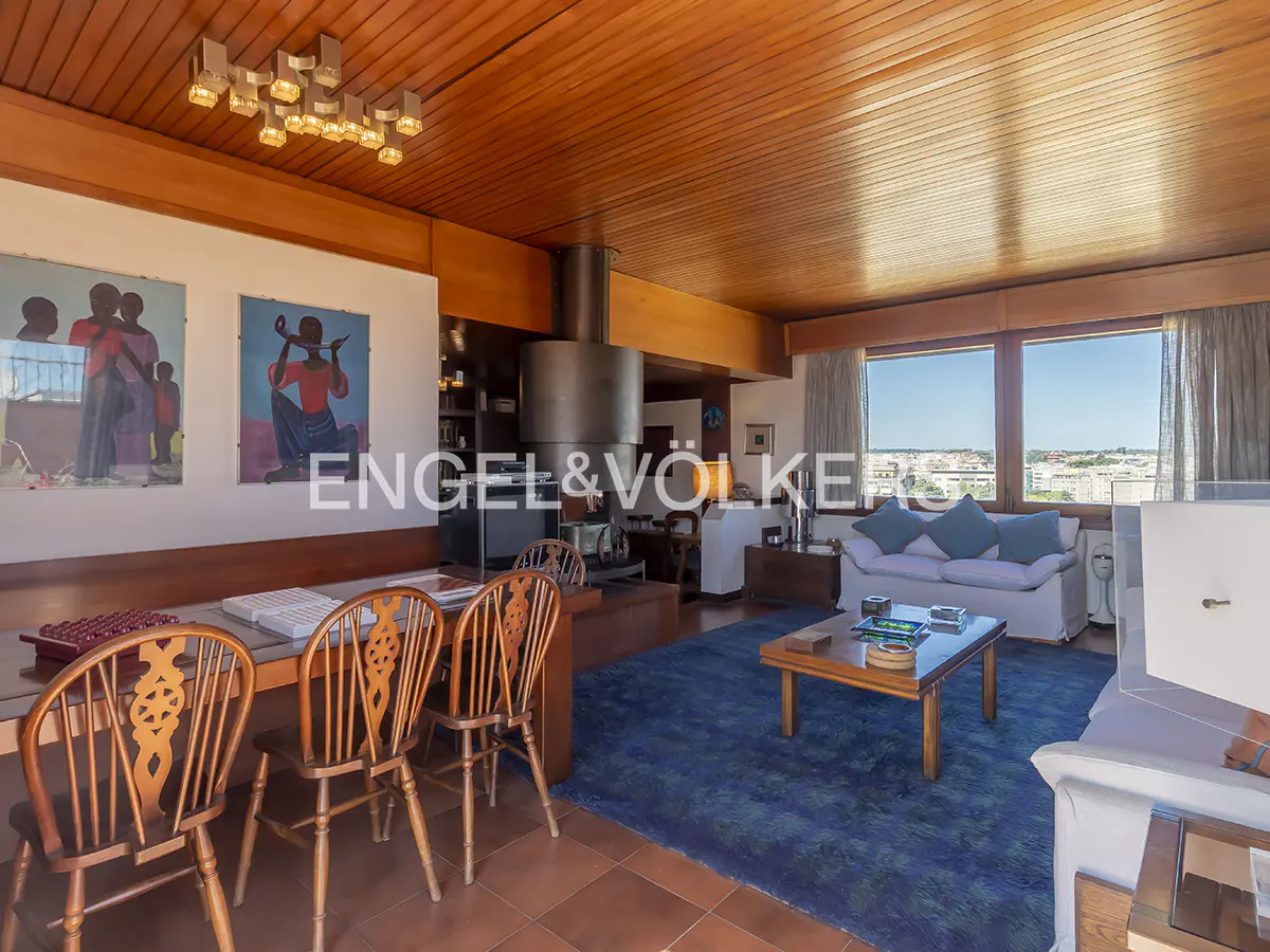A living room with wood paneling, a dining table with chairs, and a blue rug. Large windows offer a city view. Artwork adorns the walls.