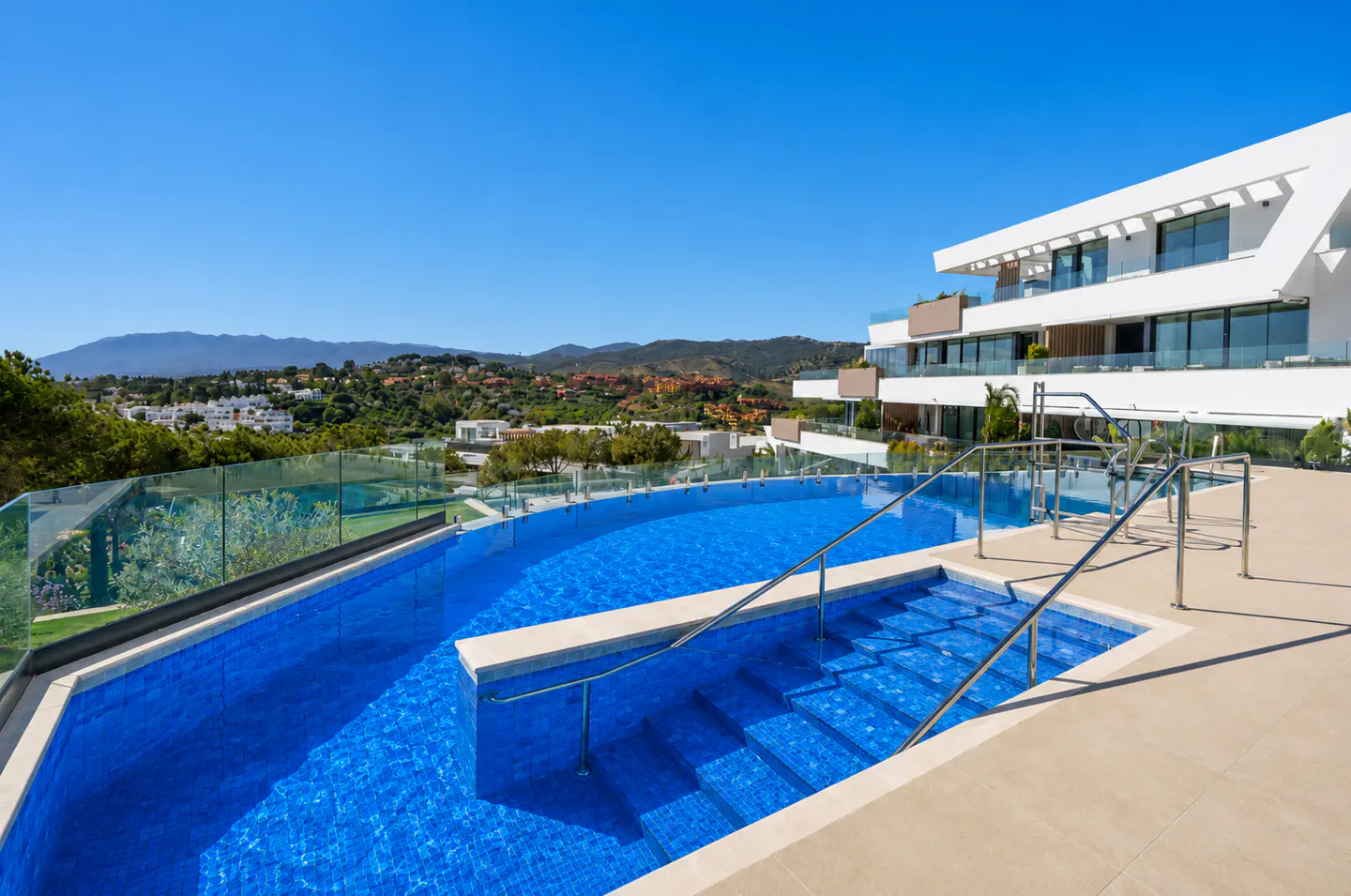 A blue tiled pool with steps and handrails, next to a modern white building under a clear blue sky.