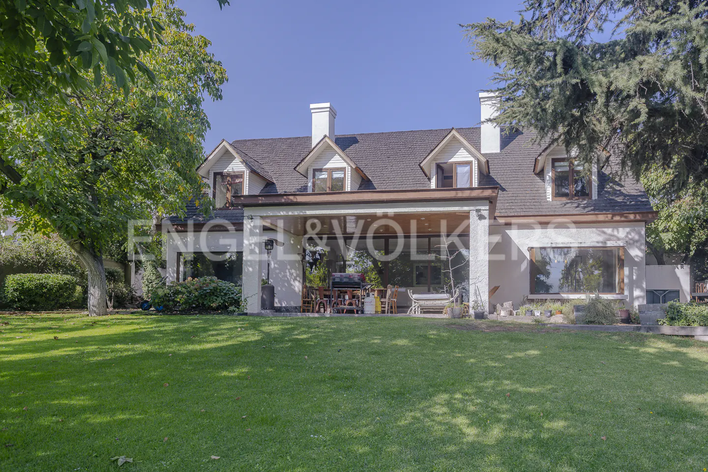 A wide shot of a two-story house with a green lawn and trees. The house has a brown roof, white walls, and a covered patio with furniture.
