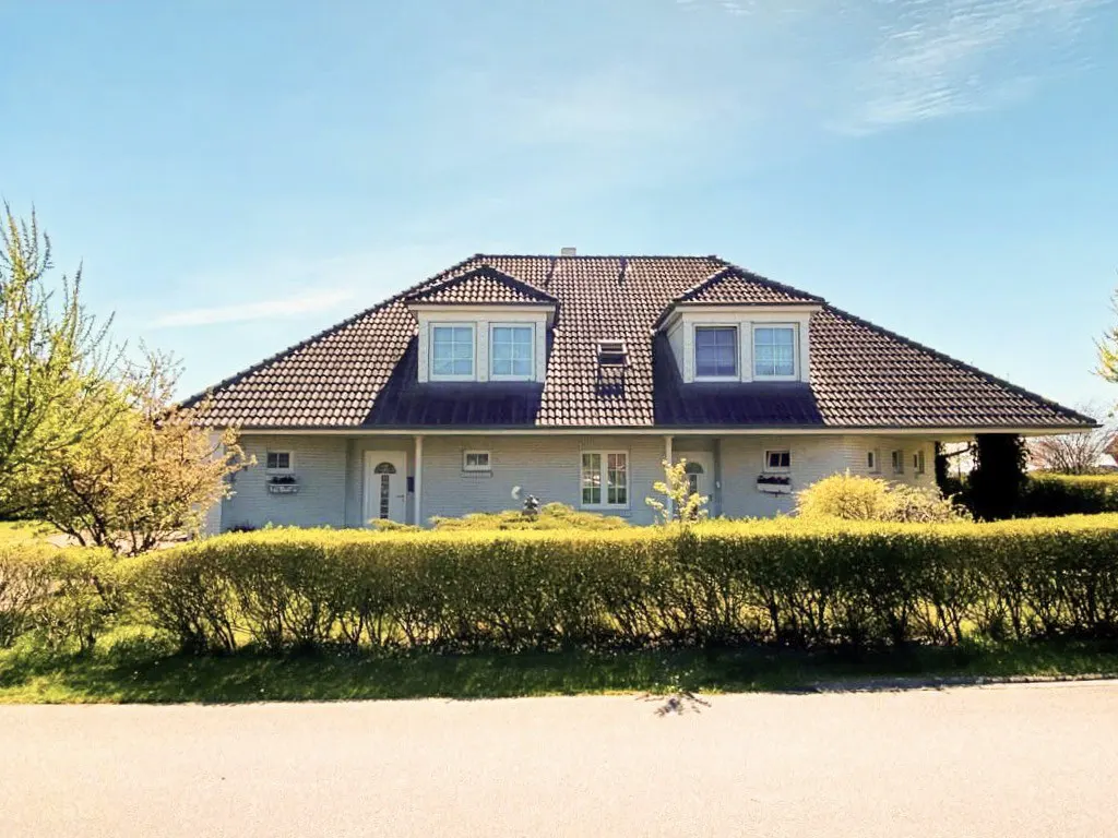A one-story light gray house with a dark brown tiled roof and dormer windows, behind a green hedge.