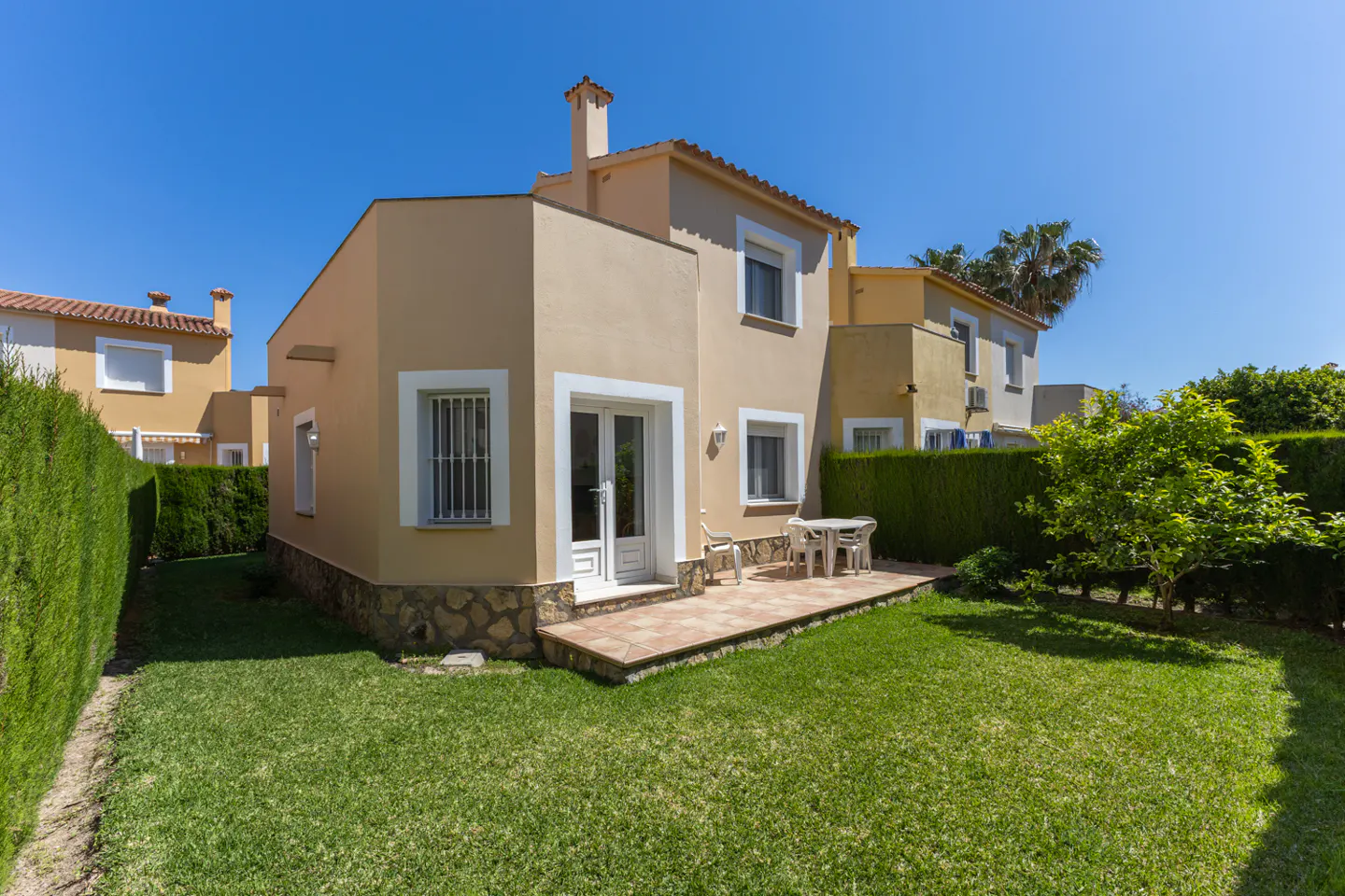Exterior view of a two-story, tan house with a green lawn, patio with table and chairs, and a blue sky.