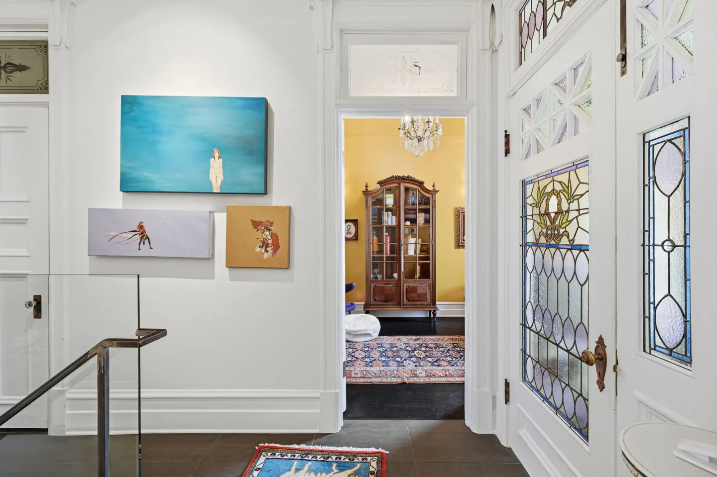 Hallway with white walls, art, and stained glass doors. A doorway leads to a yellow room with a chandelier and a wooden cabinet.