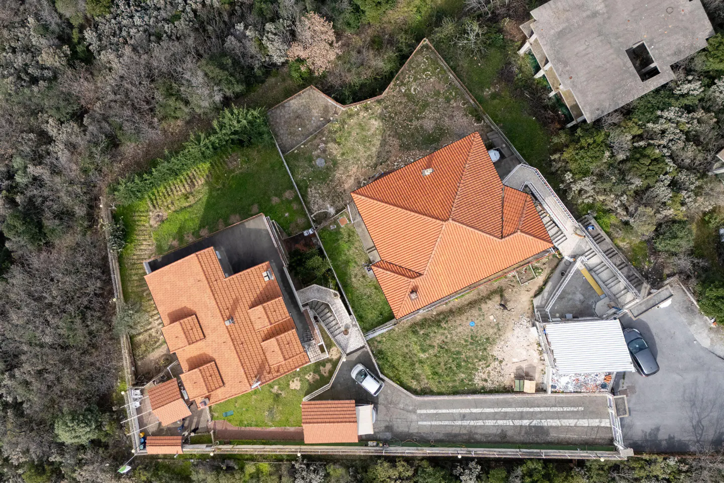 Aerial view of two houses with orange tiled roofs, surrounded by trees, driveways, and small grassy yards.