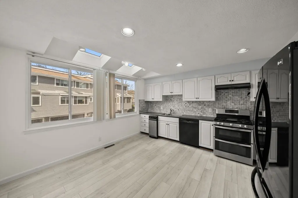 Bright kitchen with white cabinets, black appliances, and light wood floors. Large windows offer a view of neighboring houses.