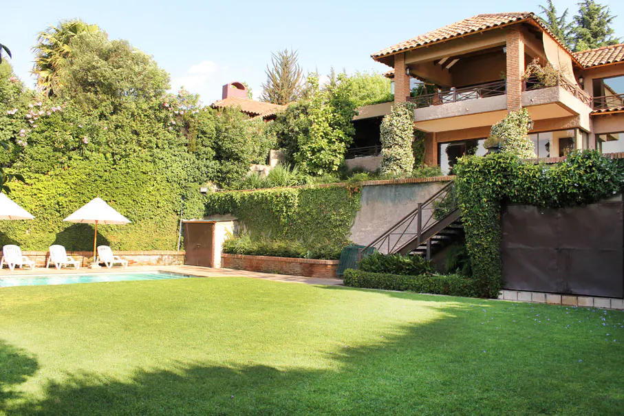 A large house with a red tile roof overlooks a green lawn, pool, and patio with white lounge chairs.