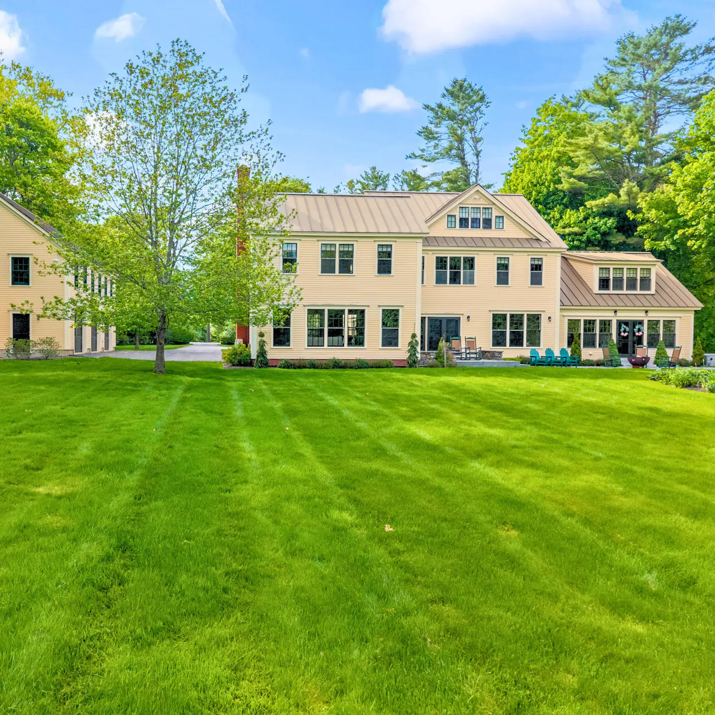 Two-story beige house with a large green lawn, trees, and blue sky.