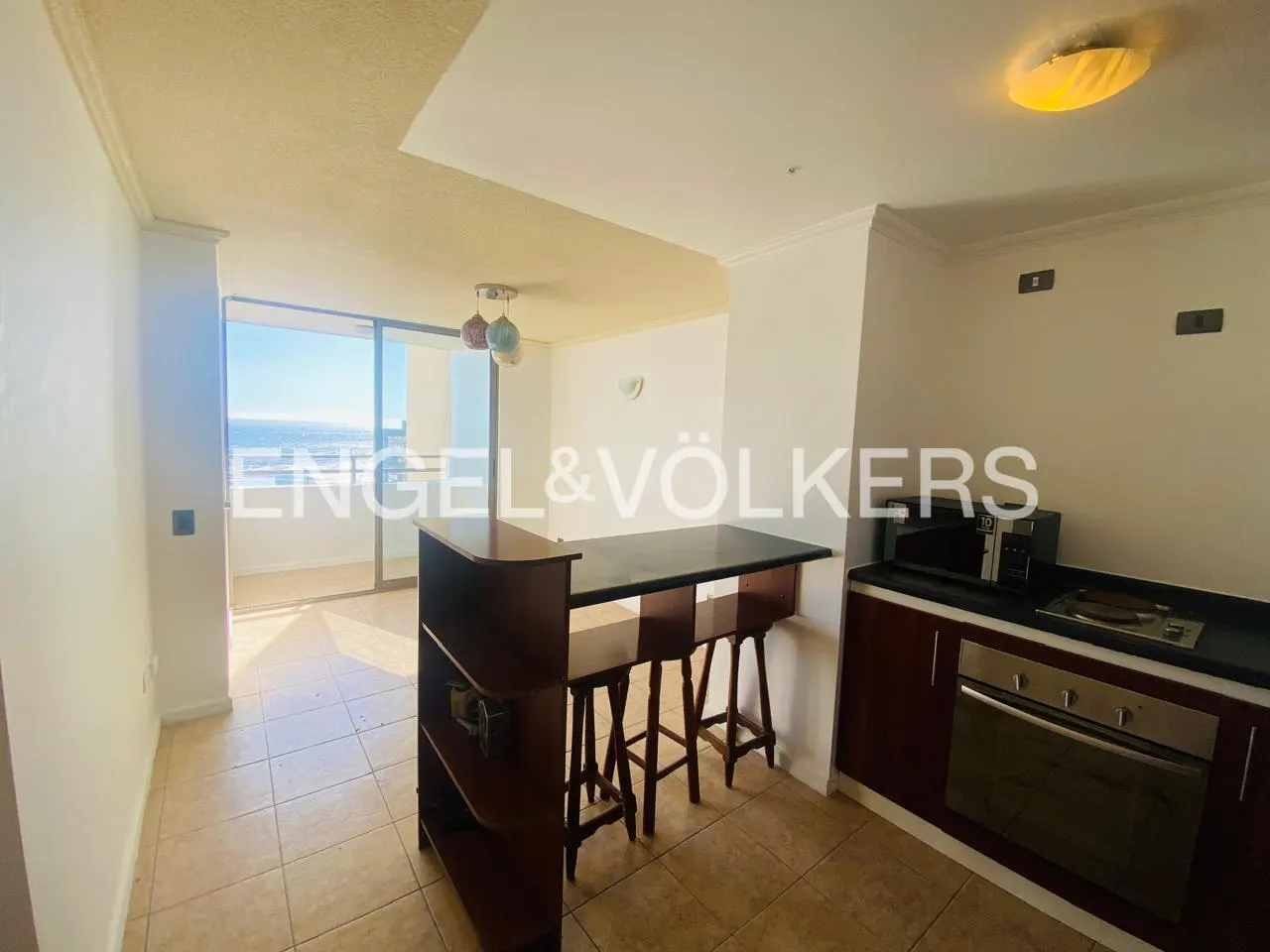 A bright kitchen with a breakfast bar, stools, and stainless steel appliances. A balcony with an ocean view is visible.