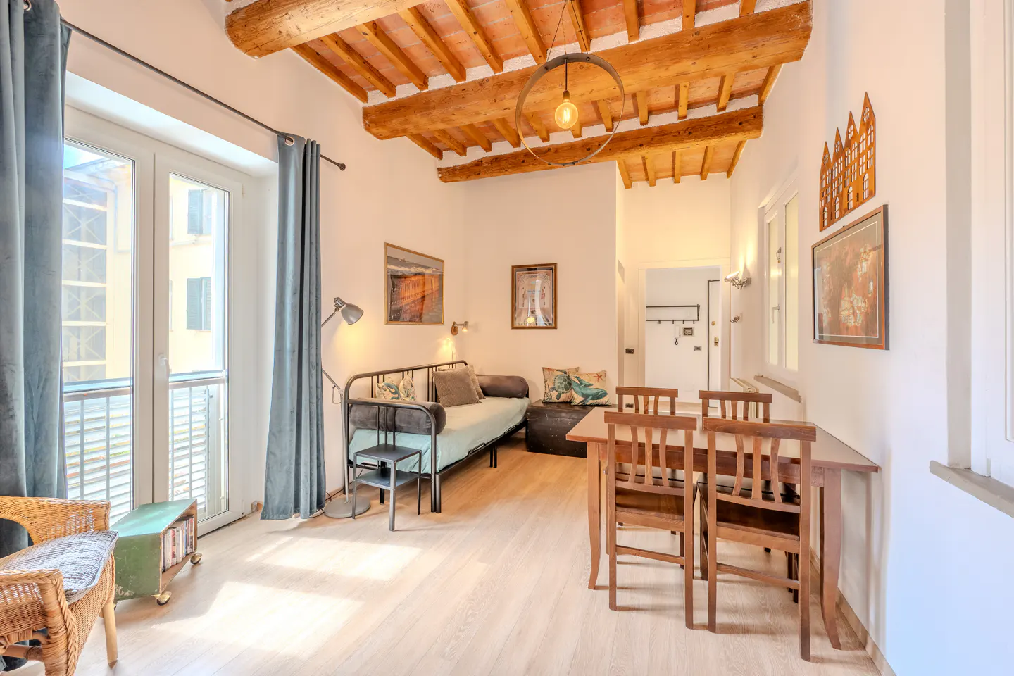 Bright living space with wood floors, exposed beam ceiling, and a dining table with four chairs. A daybed sits near a window with blue curtains.
