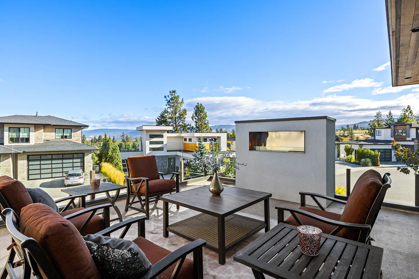 Outdoor patio with brown cushioned chairs, dark tables, and a white fireplace feature. Houses and blue sky in the background.