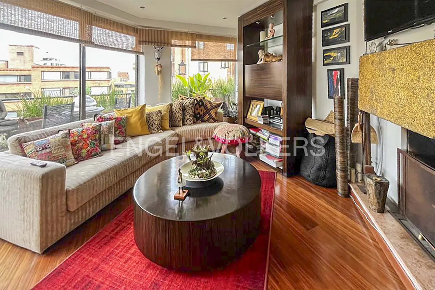 Living room with a beige sofa, colorful pillows, and a round wooden coffee table on a red rug. A bookcase and fireplace are visible.