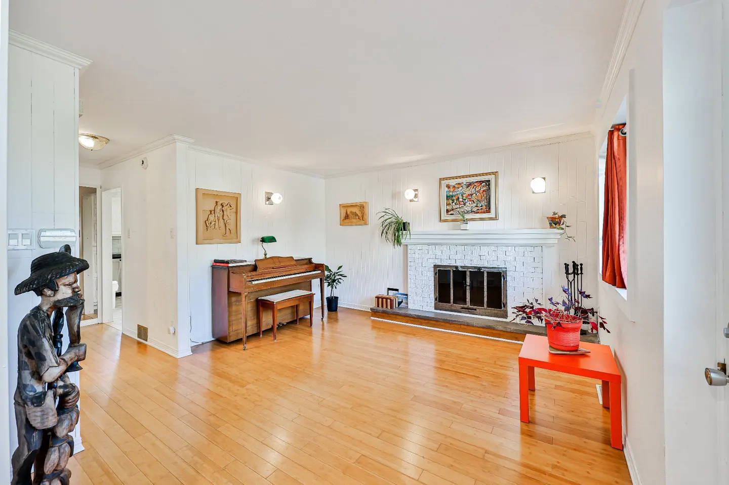 Bright living room with hardwood floors, white walls, and a brick fireplace. A piano sits near the wall, and an orange table is by the window.