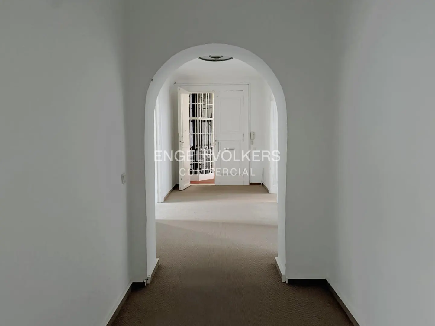 Hallway with white walls and brown floor, leading to a white door with a security gate. Archway frames the entrance.