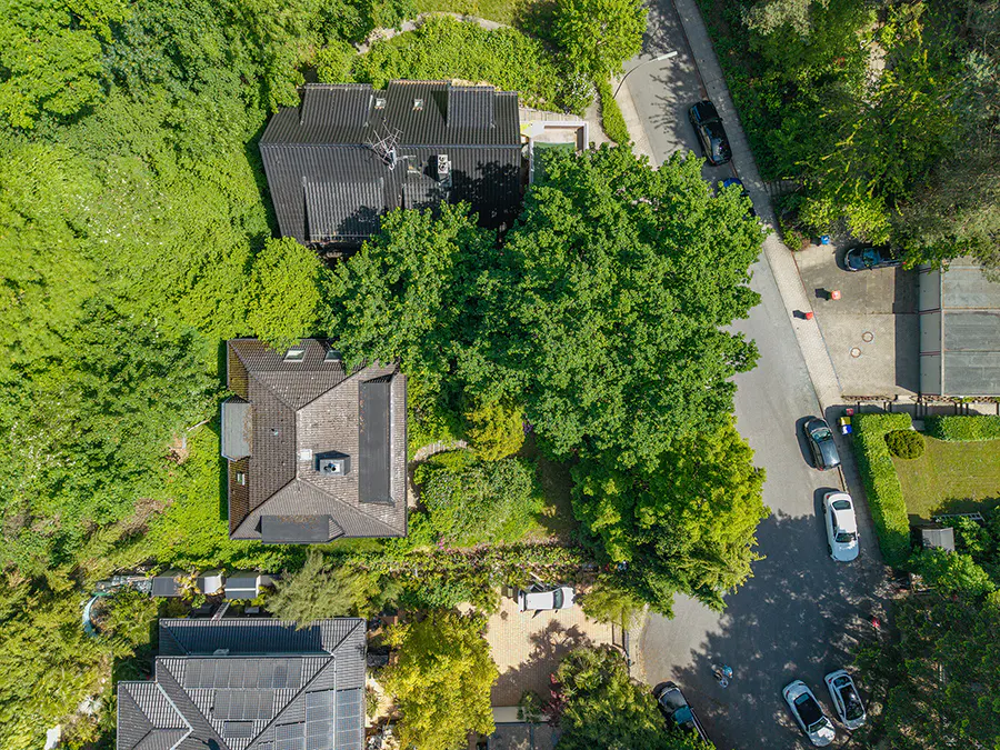 Aerial view of houses with dark roofs surrounded by green trees and a road with parked cars.