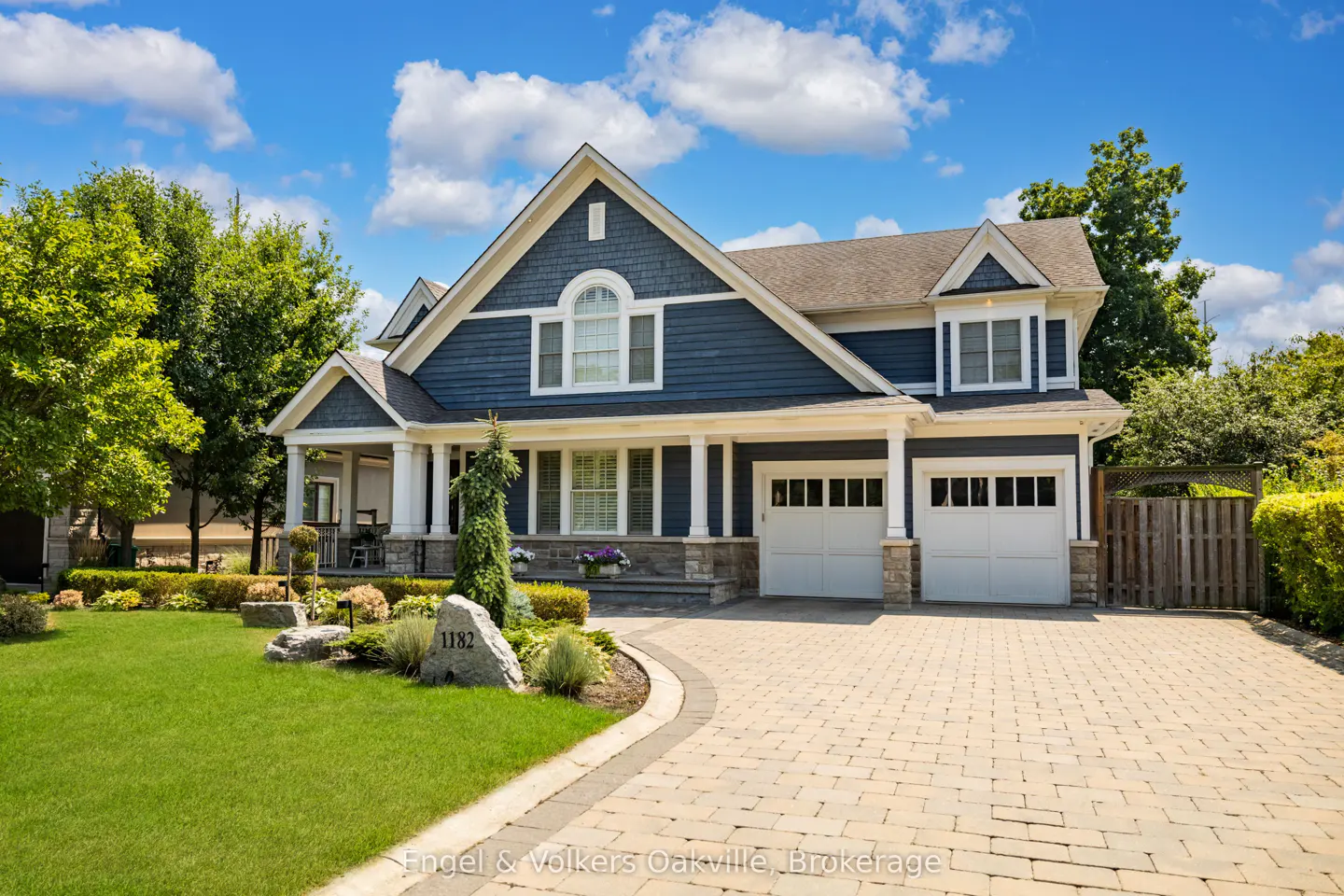 Two-story blue house with white trim, a stone facade, and a brick driveway on a sunny day.