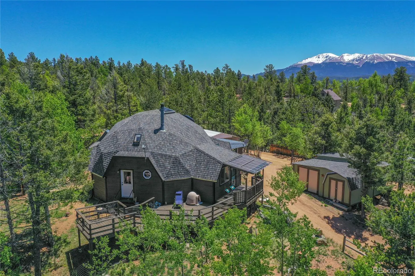 Aerial view of a dome-shaped house with a gray roof, surrounded by green trees, with snow-capped mountains in the background under a blue sky.