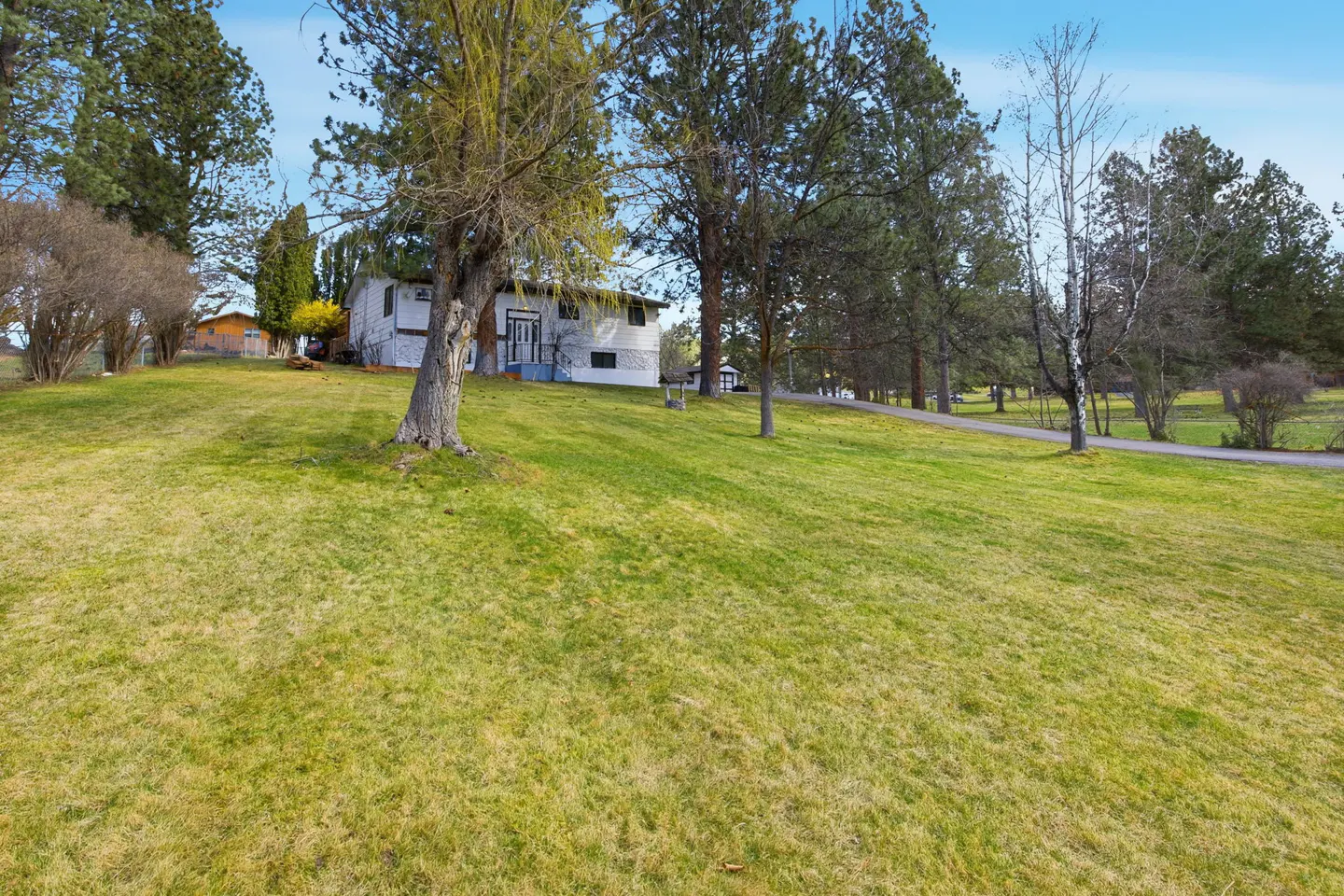 A white two-story house sits on a hill with a large green lawn and tall trees under a blue sky.