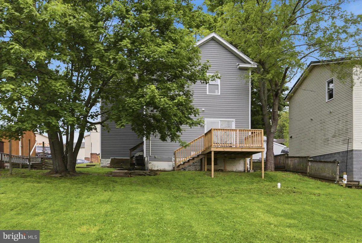 Backyard view of a two-story gray house with a wooden deck and stairs, surrounded by green trees and grass.
