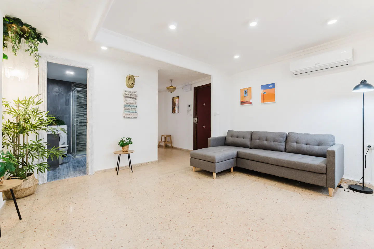 Bright living room with gray sectional sofa, terrazzo floor, and white walls. Artwork, plants, and a glimpse of a bathroom add detail.
