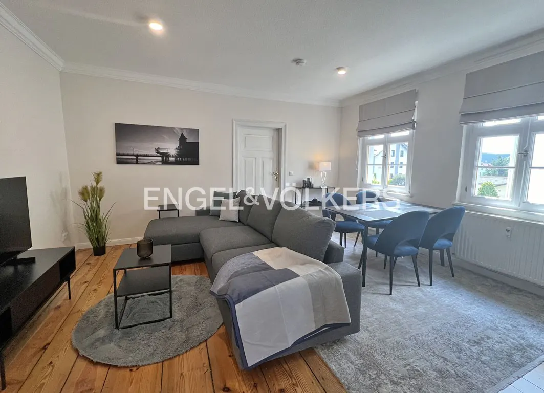 Living room with gray sectional sofa, dining table with blue chairs, and wood floors. A black and white photo hangs on the wall.