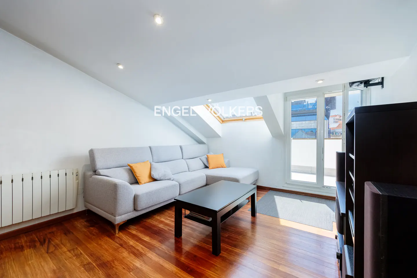 Bright living room with hardwood floors, a gray sectional sofa with orange pillows, and a black coffee table. A window and skylight provide natural light.