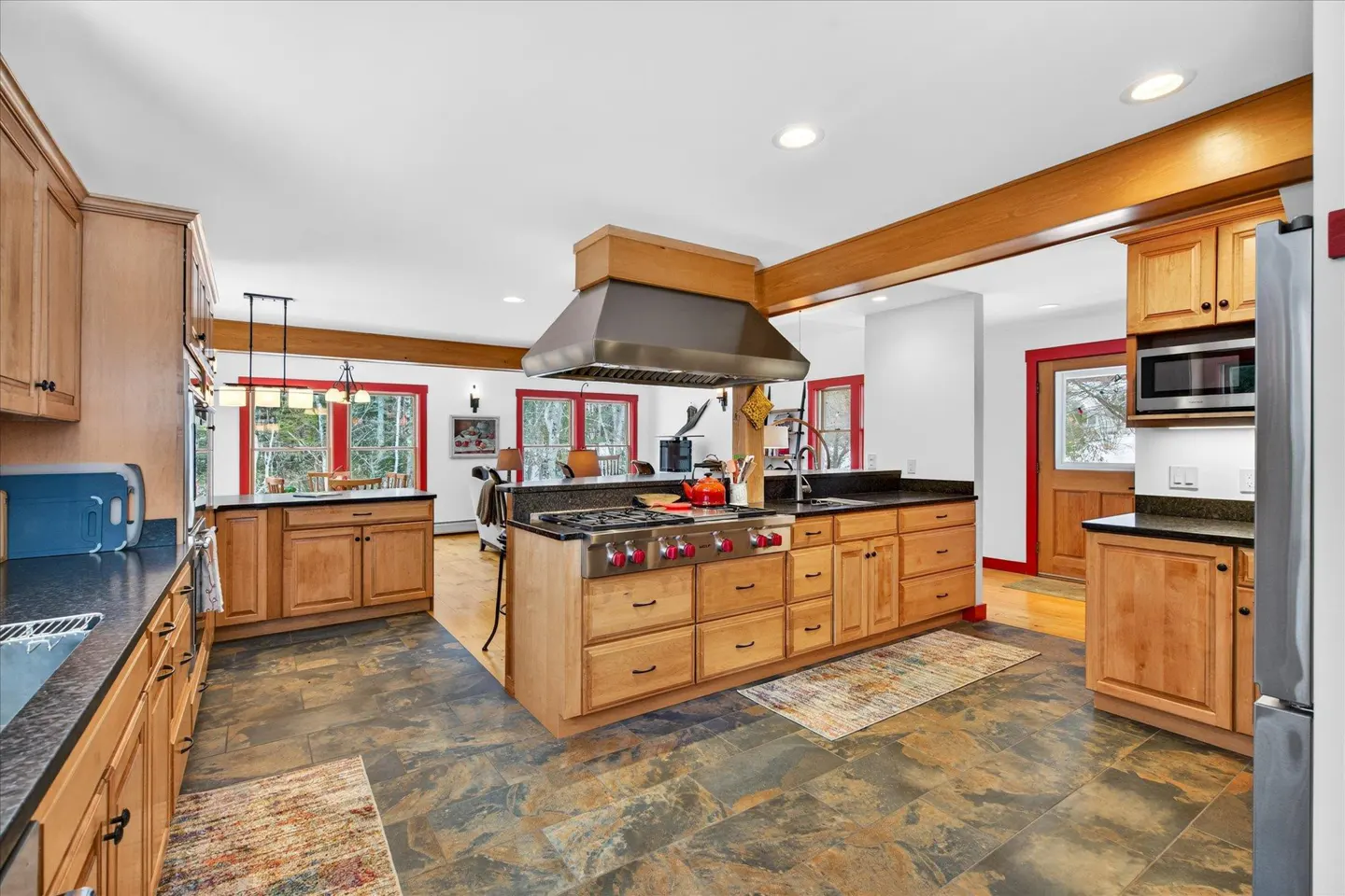 Bright kitchen with wood cabinets, black countertops, and stainless steel appliances. A large island features a cooktop and vent hood. Red window trim.