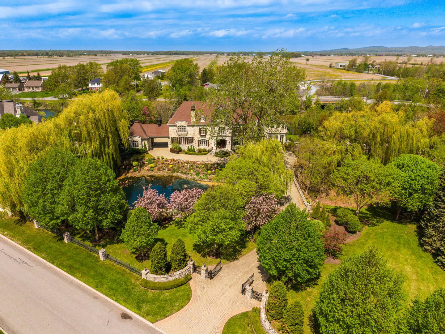 Aerial view of a large stone house with a red roof, surrounded by lush green trees and a pond. A paved driveway leads to the gated entrance.
