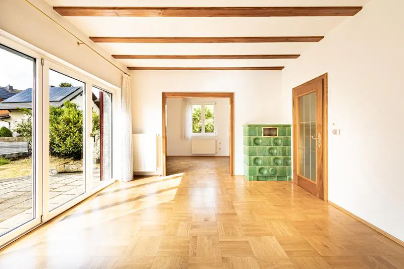 Bright room with parquet floors, exposed wooden ceiling beams, a green tiled stove, and glass doors leading to a patio.