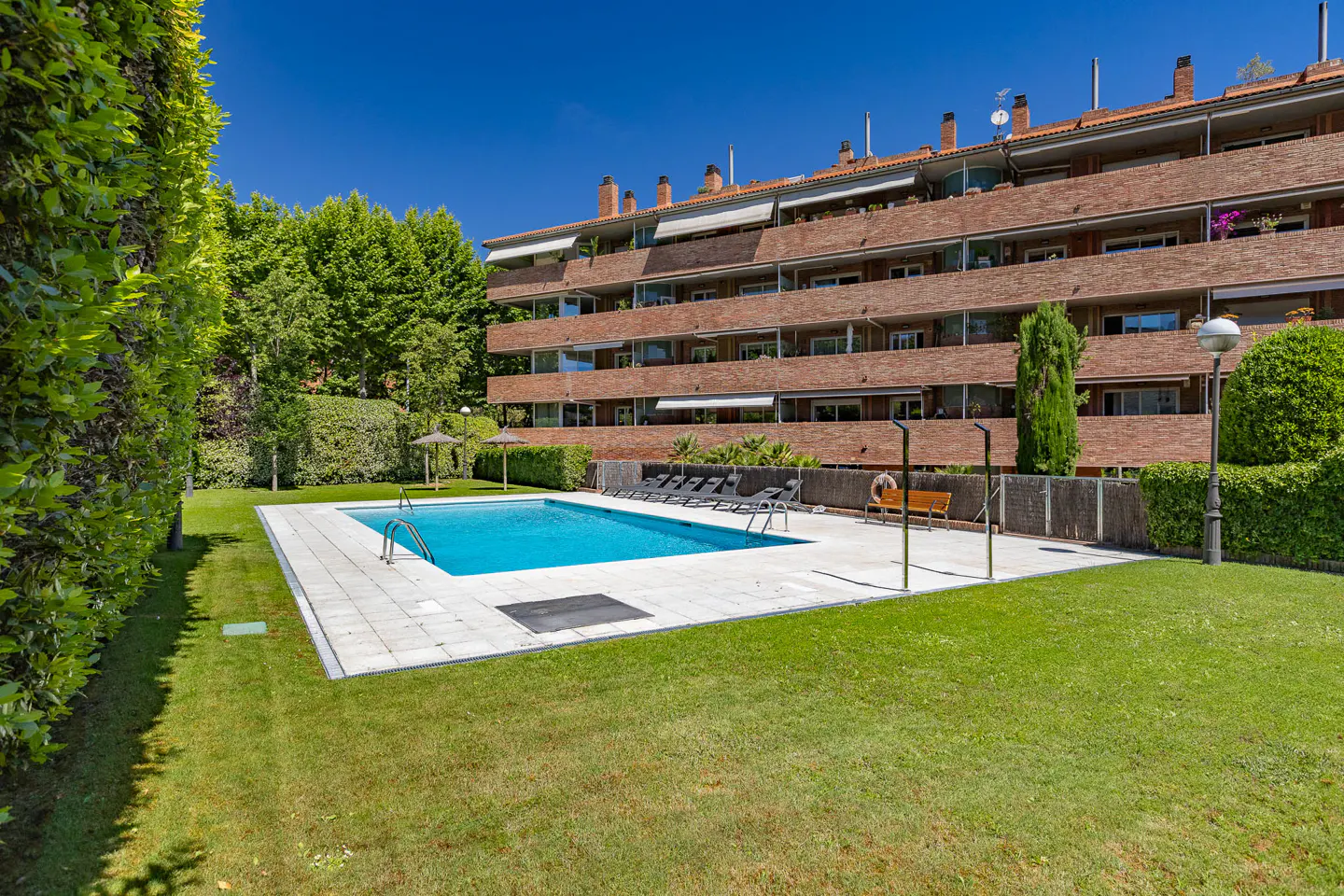 Swimming pool with blue water and lounge chairs on a patio, set in a grassy yard before a multi-story brick apartment building.