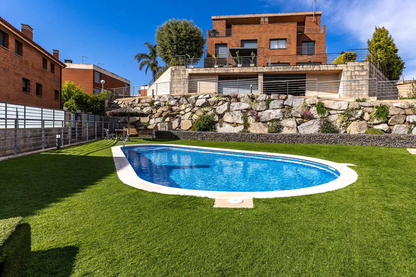 Backyard view of a blue pool surrounded by green grass, a stone wall, and a brick house under a clear blue sky.