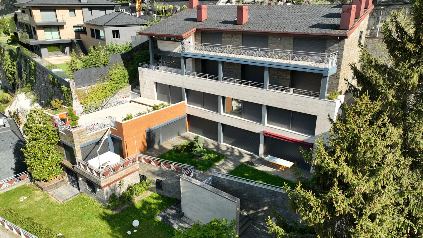 Aerial view of a multi-story stone house with balconies, dark shutters, and a terraced garden with green lawn and trees.