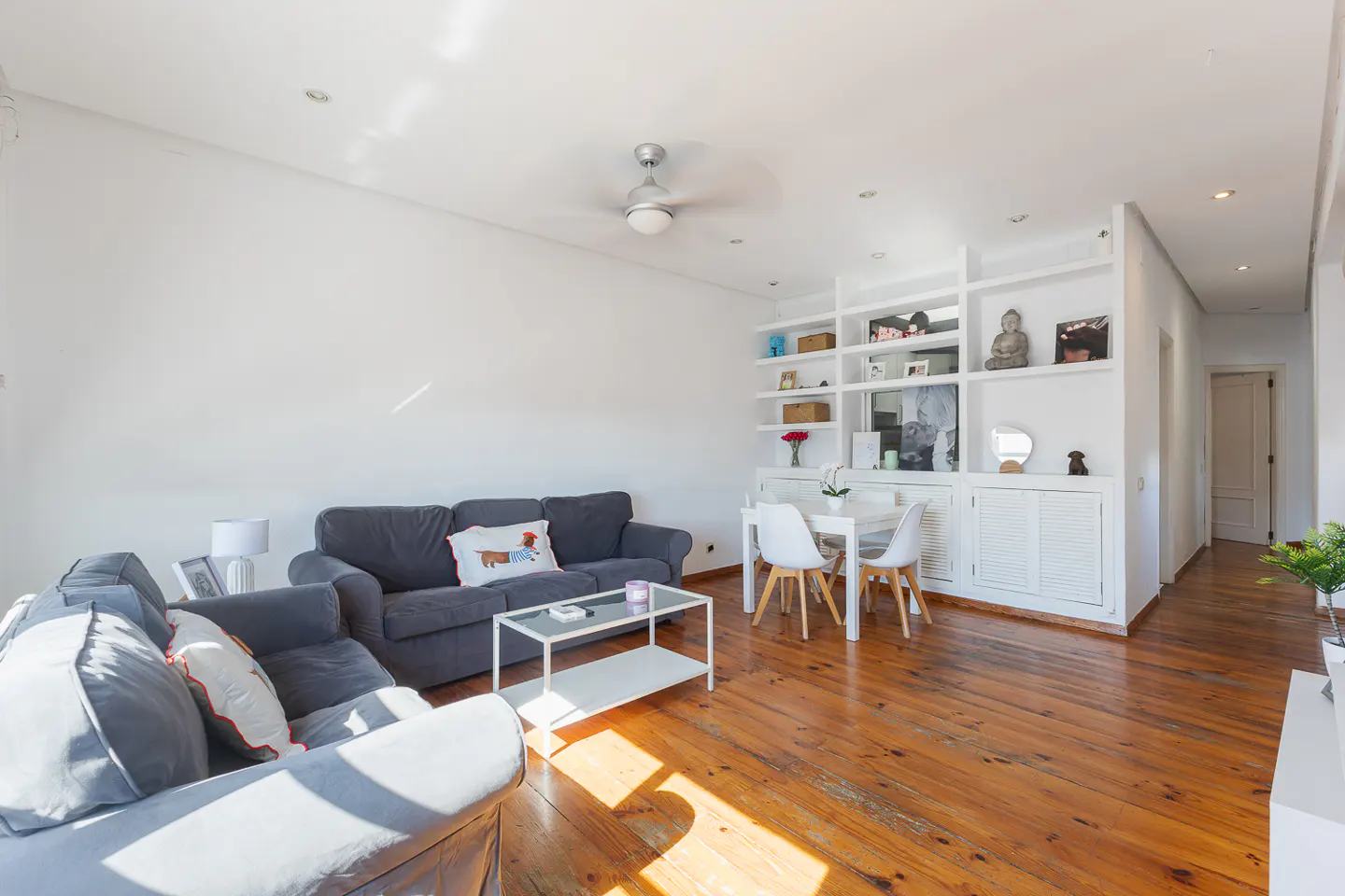 Bright living room with hardwood floors, white walls, and gray sofas. A white bookshelf and dining table are in the background.