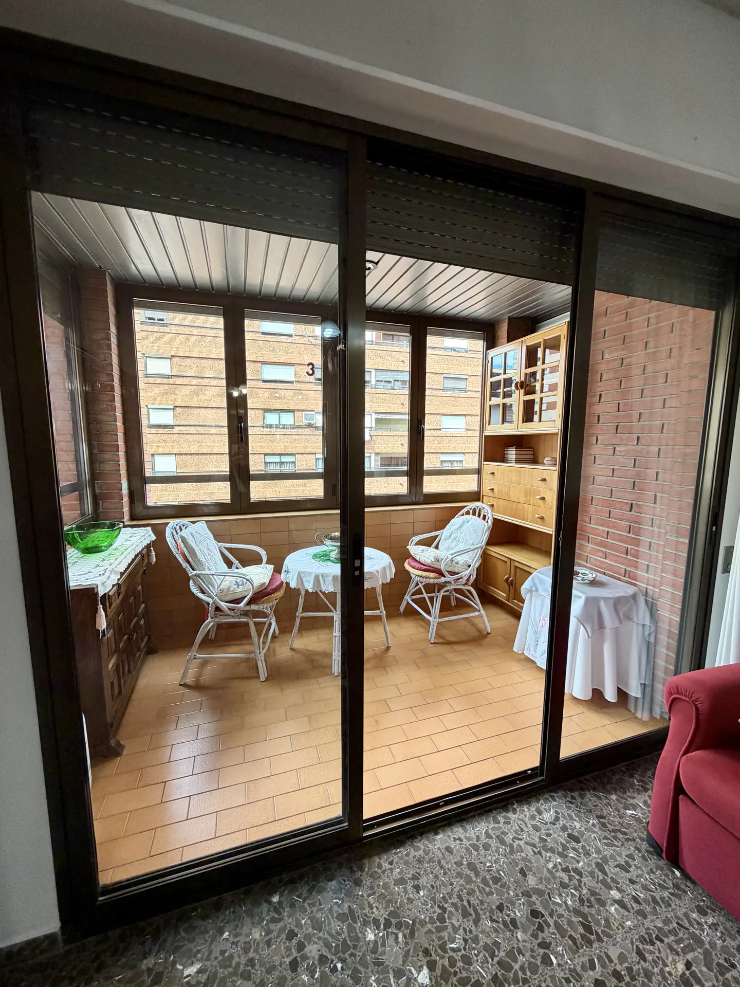 View through sliding glass doors to a sunroom with wicker chairs, small tables, and a brick wall.